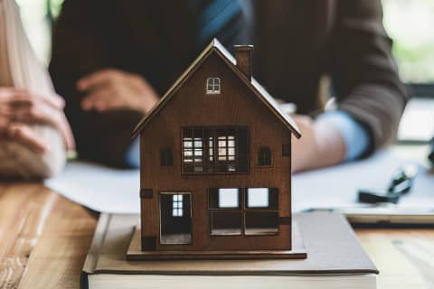 小さな木製の家の模型、後ろに契約書に署名している人物たち。