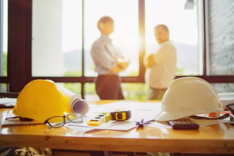 黄色と白のヘルメット、測定ツールと設計図、後ろに話している人々。